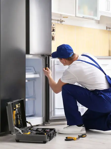 Técnico de macacão azul e boné consertando uma geladeira aberta, com a maleta de ferramentas no chão